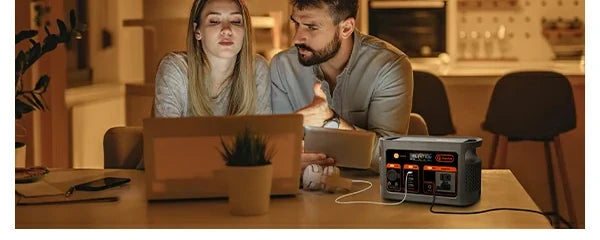 Two people with a laptop and tablet beside a FlashFish E103 portable power station on an indoor table.