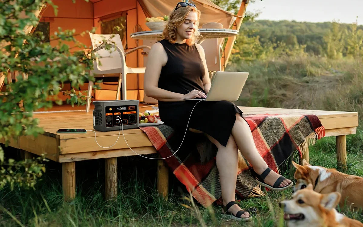 A camper uses a laptop beside a portable power station outside an orange tent, with two dogs in the foreground.