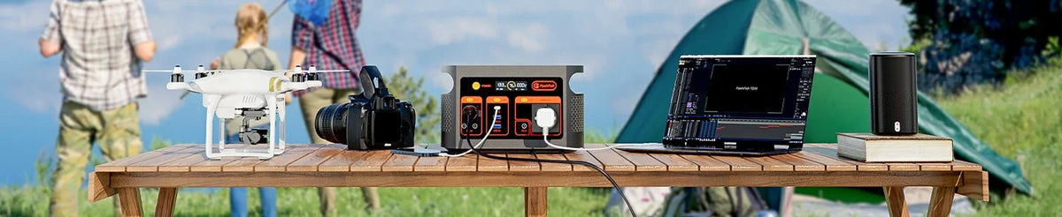 FlashFish E103 portable power station, camera, drone, laptop and speaker on an outdoor table beside a tent.