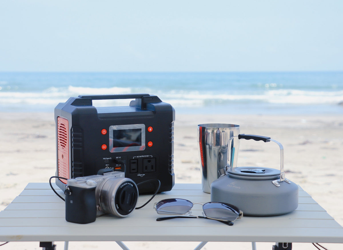 Portable power station, camera, sunglasses, metal cup and lantern on a table near the beach.