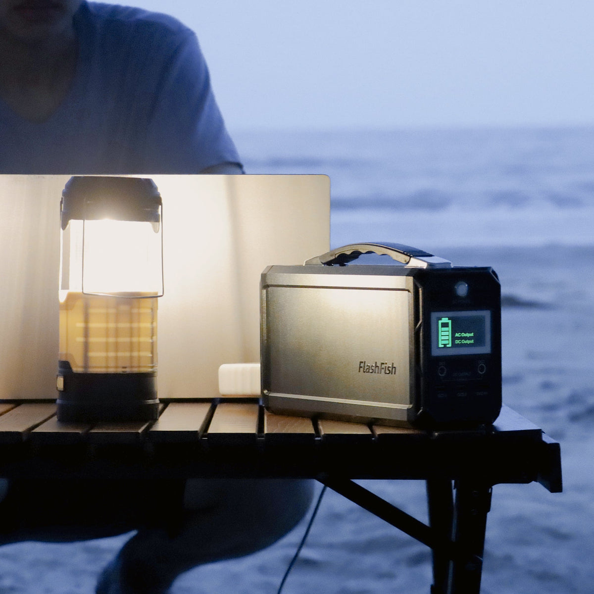 A portable power station and illuminated lantern sit beside a laptop on an outdoor table at dusk.