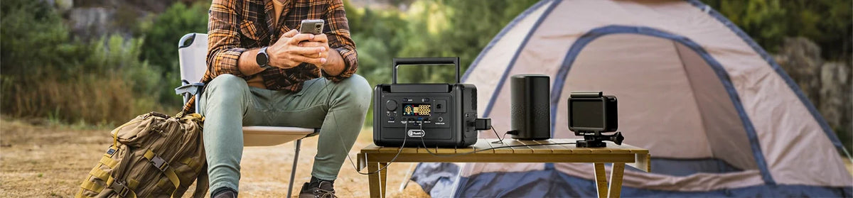 A seated camper holds a phone beside a table with a portable power station, speaker and camera.