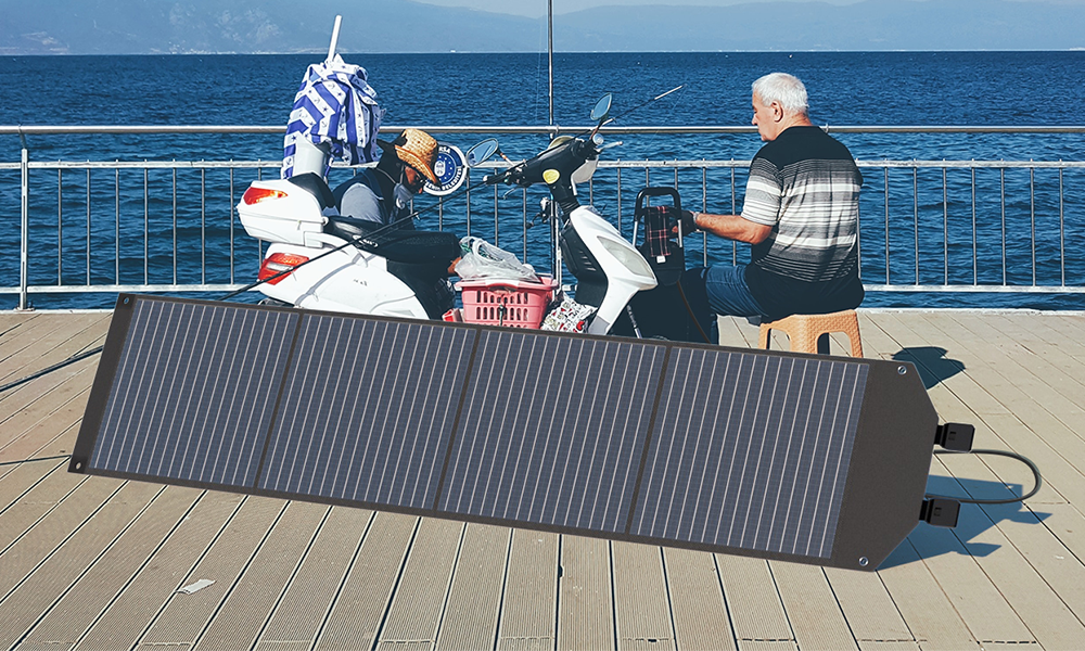 A folding solar panel stands on a wooden waterfront deck, with a seated person and scooters behind it.