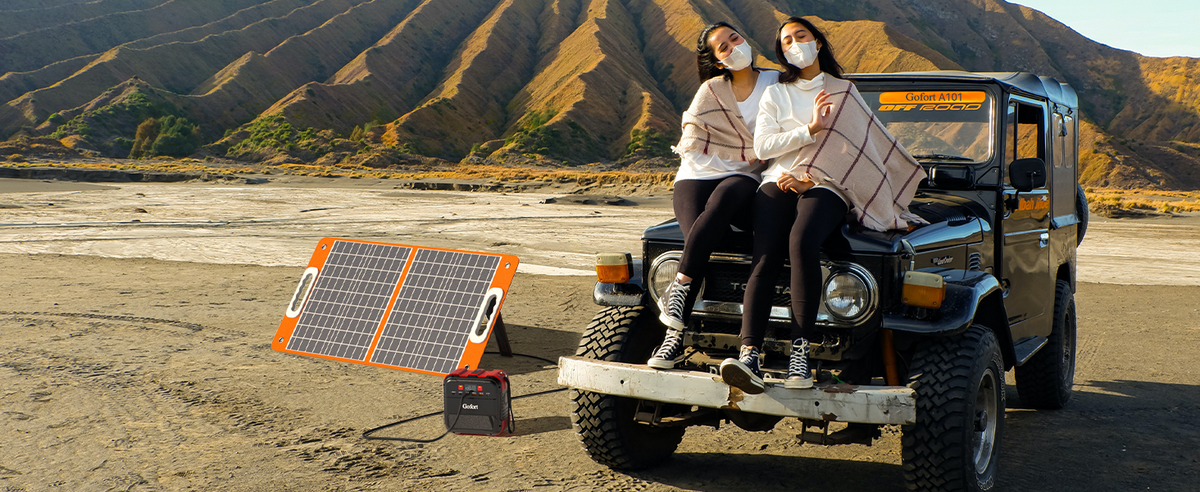 Two people sit on an off-road vehicle beside a portable power station and an unfolded solar panel.