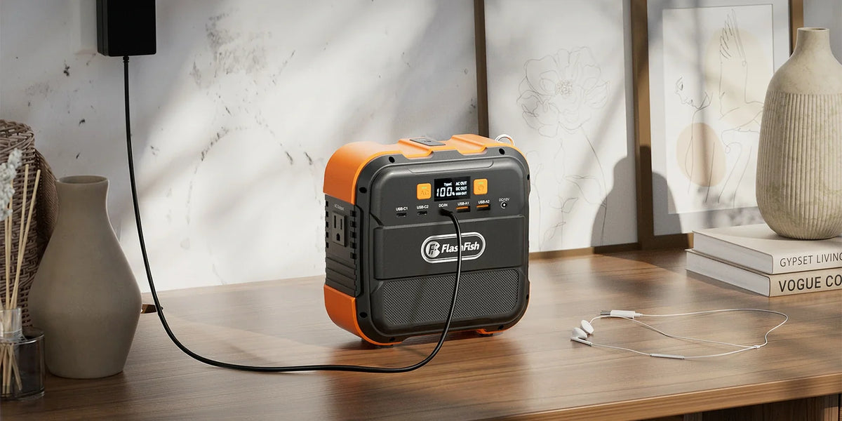FlashFish A101 portable power station connected to a wall adapter on a wooden desk beside earphones and framed art.