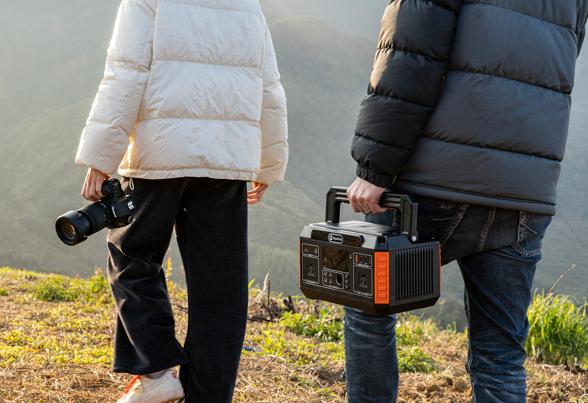 Two people stand outdoors; one holds a camera and the other carries a portable power station.