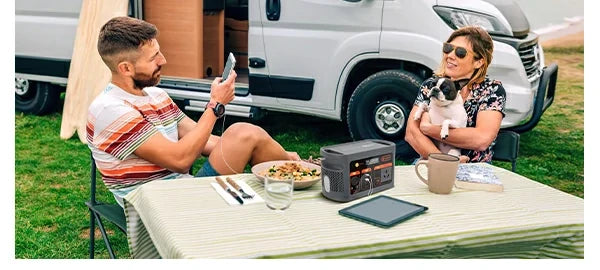 Portable power station and tablet on a campsite table beside people and an RV
