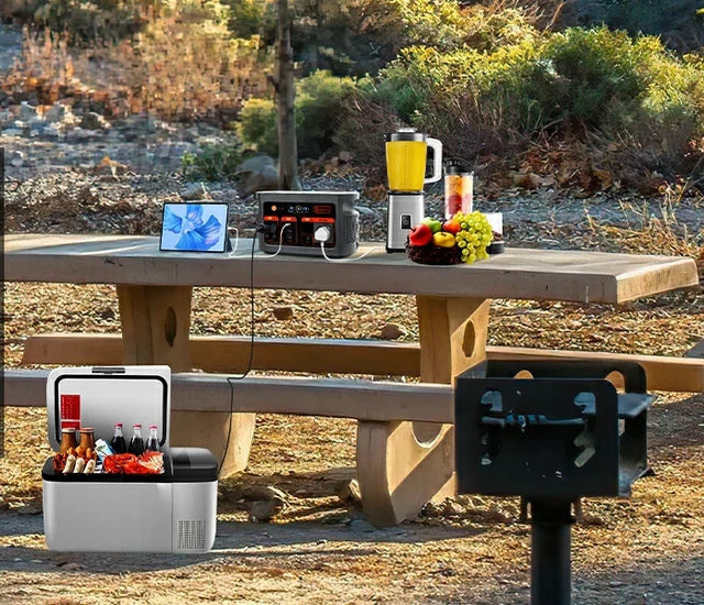 A portable power station, tablet and blender on a picnic table, with a cooler and grill nearby.